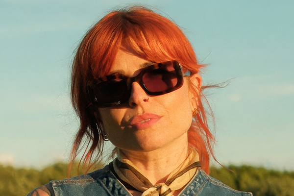 A lady with ginger hair tied back, wearing sunglasses and a short-sleeved denim jacket.