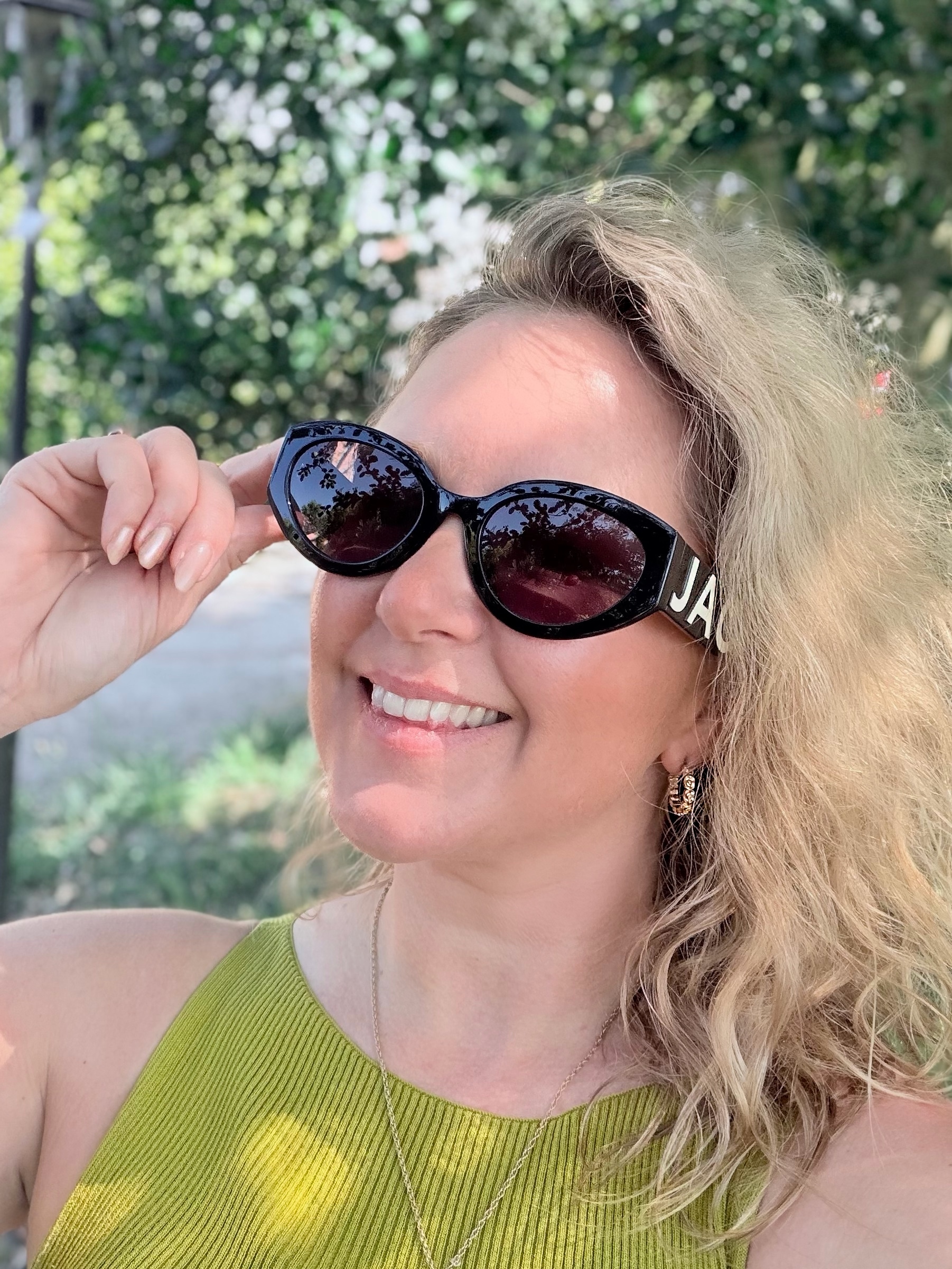 Lady with blond hair and a green top smiles as she wears a pair of Marc Jacobs sunglasses.
