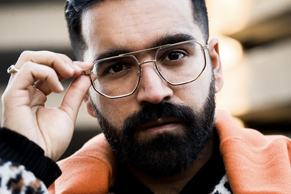 A man with black hair and a beard wearing metal framed glasses and an orange jacket.