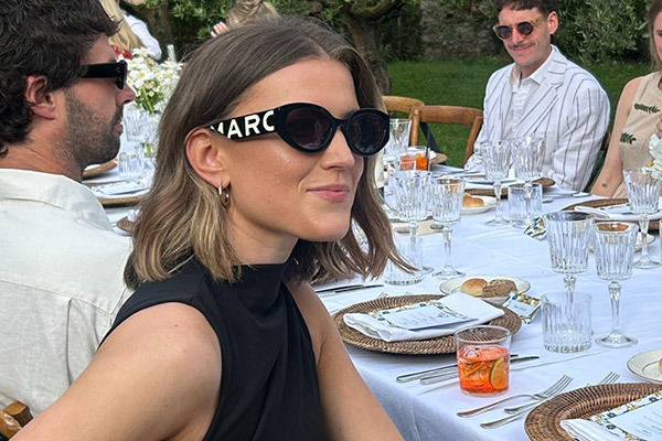 Woman in black dress sitting at a table in a garden wearing black Marc Jacobs sunglesses